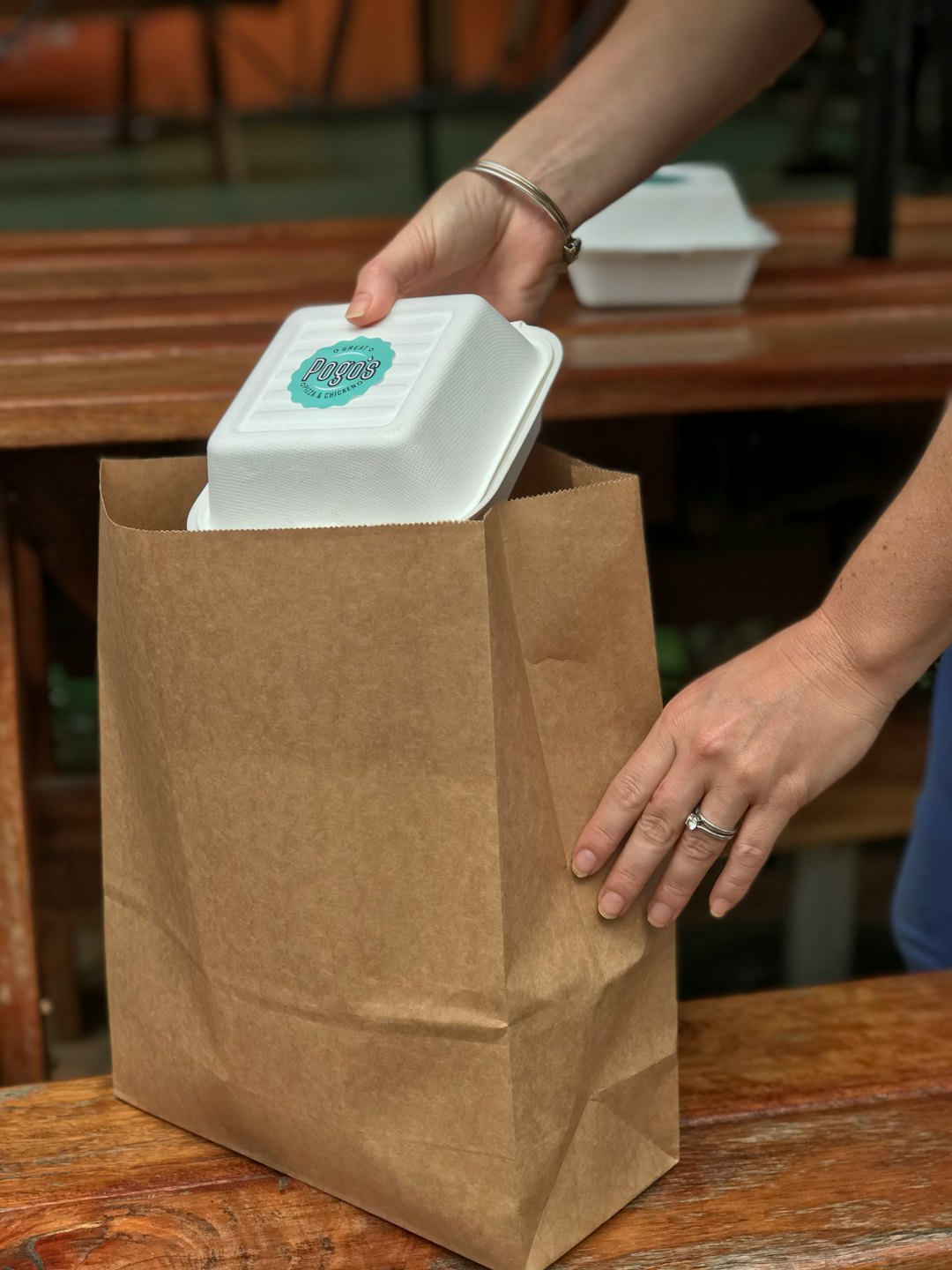 Hands place takeout container into brown paper bag on table.